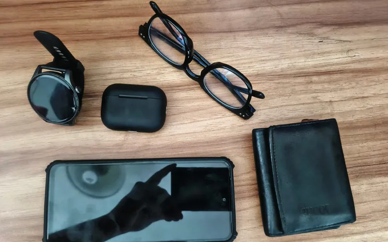A wallet, a phone, a pair of glasses, a watch and an earbud case laid out on a wooden table.