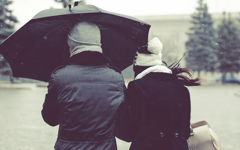 Two people seen from behind walking close together under one black umbrella in falling snow, in winter coats and knitted hats.