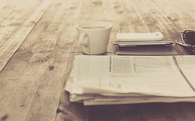 A white cup of coffee, a phone lying face down beside a pen, and a folded stack of newspapers on a bare wooden table in daylight.