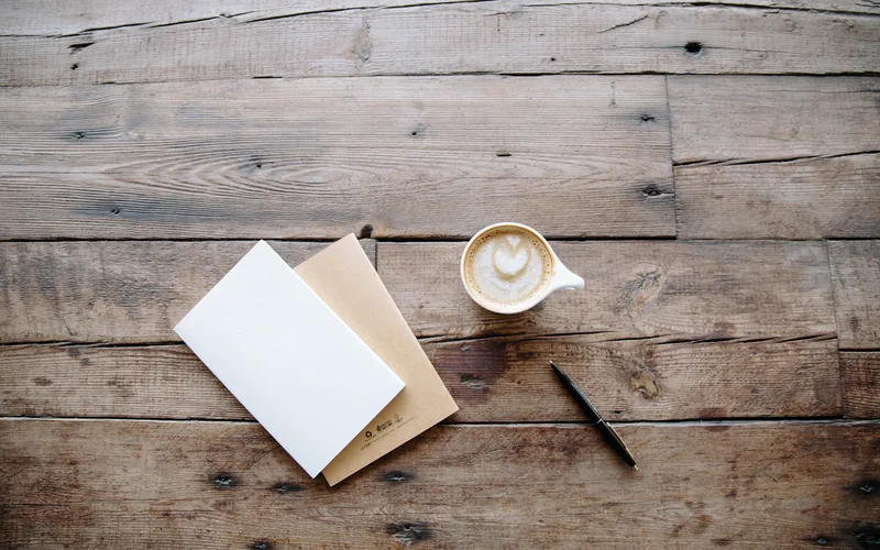 A bare wooden table seen from above, with a white card squared on top of a kraft notebook, a pen beside them and a cup of coffee.