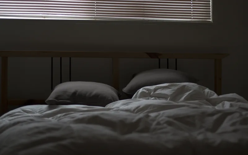 An empty unmade bed with the duvet pushed aside and two grey pillows, in a dim room lit only by pale light coming through the slats of a closed blind above the headboard.