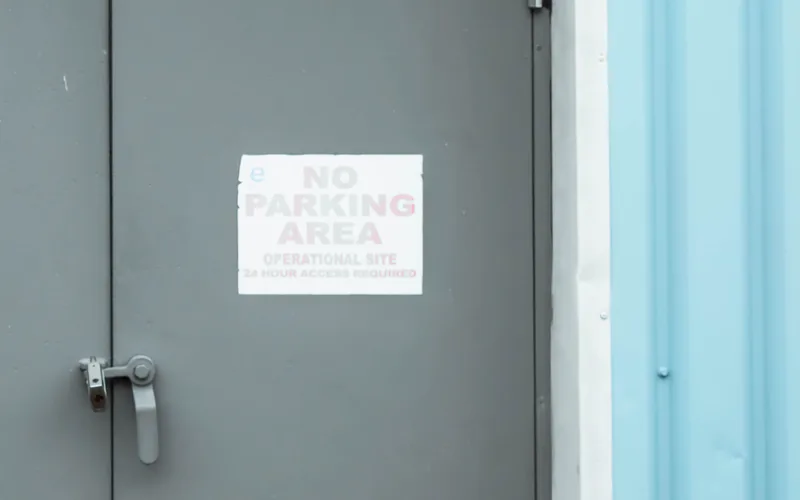 A grey metal service door with a small printed notice beside its lever handle, set into a pale blue corrugated wall in plain daylight.